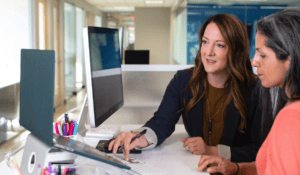 Two professional women collaborating at a white office desk, looking together at a desktop computer monitor while working on a laptop.