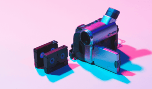 A vintage silver miniDV camcorder with its flip-out LCD screen open, set against a pink background with neon blue and pink lighting, alongside two small black MiniDV video cassettes standing upright.