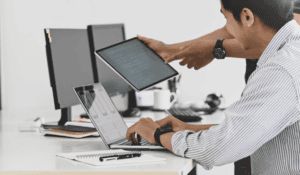 Two IT consultants collaborating at a desk with a laptop, tablet, and desktop monitors.