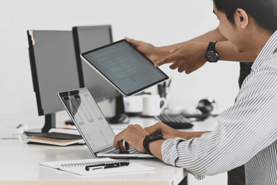 Two IT consultants collaborating at a desk with a laptop, tablet, and desktop monitors.