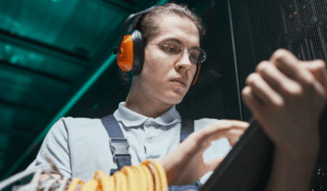 A technician wearing orange safety earmuffs and glasses updates records on a digital tablet in a server room.