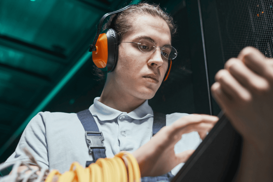 A technician wearing orange safety earmuffs and glasses updates records on a digital tablet in a server room.