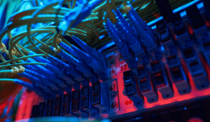 Close-up view of multiple fiber optic cables with blue connectors plugged into a numbered patch panel inside a server rack, illuminated by red and blue ambient lighting.