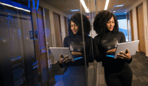 An IT professional with curly hair stands in a modern data center hallway next to a glass wall, typing on a white tablet computer. Behind her, server racks glow with blue lights inside a server room.
