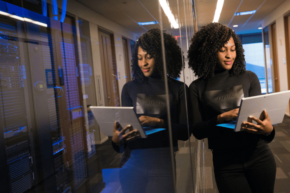 An IT professional with curly hair stands in a modern data center hallway next to a glass wall, typing on a white tablet computer. Behind her, server racks glow with blue lights inside a server room.