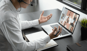 A business professional wearing a headset participates in a remote video conference call on a laptop while taking notes on a desk.