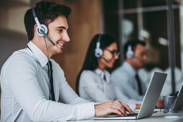 A smiling male office worker wearing a headset and tie sits at a desk typing on a laptop, with two blurred colleagues in the background also wearing headsets.
